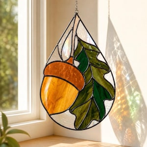 May include: A teardrop-shaped stained glass suncatcher featuring an amber acorn with a dark orange cap and a textured green oak leaf. The piece is framed in dark metal solder and hangs from a silver chain, casting colorful light reflections.