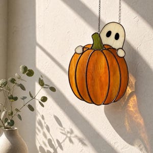 May include: A stained glass suncatcher featuring an orange pumpkin with a green stem and a small white ghost peeking out from behind it. The piece is constructed with dark metal soldering lines and hangs from a thin metal chain for display.