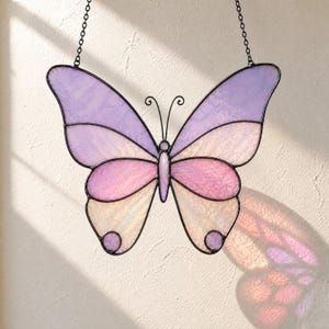 May include: A stained glass butterfly suncatcher featuring pastel purple, pink, and cream-colored glass panels held together by dark metal soldering. The butterfly has two thin metal antennae and is suspended by two black metal chains for hanging.