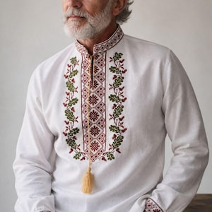 Op de afbeelding: Een wit shirt met lange mouwen, een opstaande kraag en een centraal verticaal paneel met ingewikkeld rood geometrisch borduurwerk. Groene eikenblad- en eikelmotieven flankeren het midden, met bijpassend borduurwerk op de manchetten en een beige kwastje.