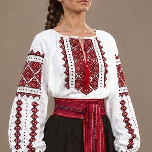 Peut inclure: Un chemisier blanc à manches longues orné de broderies géométriques rouges et noires sur la poitrine et les manches, avec un col à cordon et pompons rouges. Une large ceinture tissée rouge et bordeaux est nouée sur une jupe noire.
