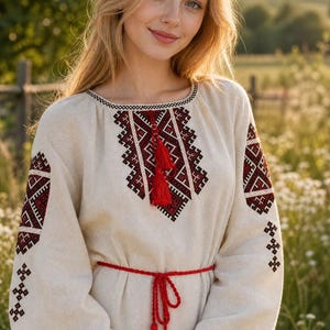 Può includere: Una camicetta beige a maniche lunghe con intricati ricami geometrici in filo rosso scuro e nero attorno allo scollo e alle maniche. Il capo presenta uno scollo rotondo con un cordoncino con nappe rosse e una sottile cintura intrecciata rossa.