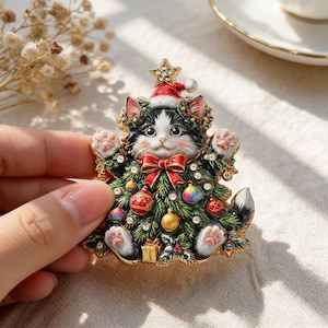 May include: A gold-toned metal brooch featuring a black and white cat wearing a red Santa hat with a star on top. The cat is surrounded by green pine branches, red bows, colourful ornaments, and clear crystals, with its paws raised upwards.