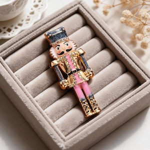 May include: A nutcracker brooch featuring a gold-tone metal finish with pink enamel accents on the jacket and legs. The figure is decorated with clear rhinestones and has a dark blue glittery hat. The brooch has a detailed face with a mustache.