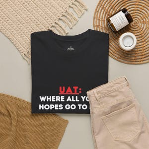 May include: A black short-sleeved t-shirt featuring the red text UAT: and white text WHERE ALL YOUR HOPES GO TO DIE. The shirt has a crew neckline and a small brand logo printed on the inside neck label area, displayed on a flat surface.
