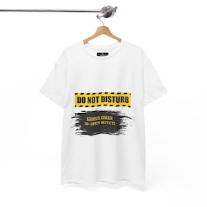 Könnte beinhalten: Ein weißes Kurzarm-T-Shirt mit einer Grafik aus gelb-schwarzen Warnstreifen und dem Text DO NOT DISTURB. Darunter befindet sich ein schwarzer Pinselstrich mit dem gelben Text KNOWN ISSUES: 50+ OPEN DEFECTS.