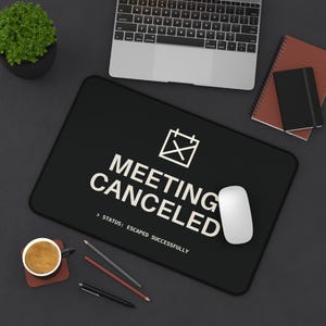 Puede incluir: Una alfombrilla de escritorio rectangular negra con un icono de calendario blanco con una X, el texto "MEETING CANCELED" y el subtítulo "STATUS: ESCAPED SUCCESSFULLY" impresos en una fuente sans-serif centrada en la superficie.