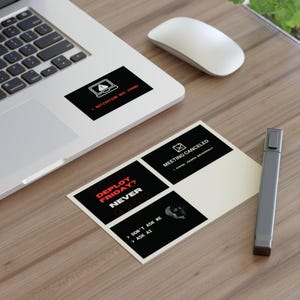 May include: A silver laptop with a black sticker featuring a laptop icon and the text MOTIVATION NOT FOUND. Next to it is a rectangular sheet of stickers with black backgrounds and white or red text including MEETING CANCELED, DEPLOY FRIDAY? NEVER, and DON'T ASK ME ASK AI.