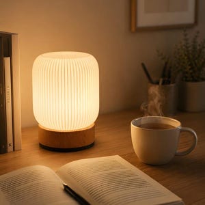 Puede incluir: Una lámpara de mesa cilíndrica con una pantalla de cristal blanco de costillas verticales y una base circular de madera clara. La lámpara emite un brillo cálido y suave, iluminando el área circundante con una luz tenue y acogedora.