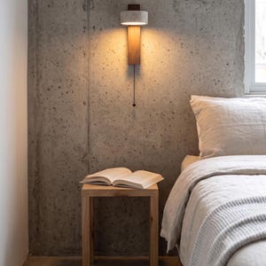 May include: A wall-mounted light fixture featuring a cylindrical white concrete shade atop a vertical rectangular wooden panel. A black pull chain hangs from the center of the fixture, which emits a warm glow against the textured concrete wall.