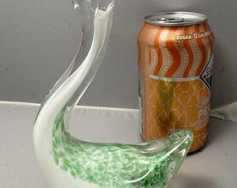 May include: A clear glass swan figurine with a long, curved neck and a white and green speckled body. The swan is next to an orange can with text.