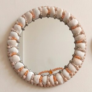 Könnte beinhalten: Ein runder Wandspiegel mit einem Rahmen, der mit einem sich wiederholenden Muster aus weißen Muscheln verziert ist, durchsetzt mit geflochtener beiger Kordel und kleinen grauen Muscheln. Ein einzelnes orangefarbenes, sternförmiges Element ist unten mittig platziert.
