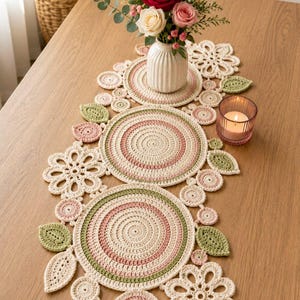 Peut inclure: Un chemin de table en crochet composé de napperons circulaires, de motifs floraux et de feuilles en fil crème, rose pâle et vert sauge. Le chemin de table est disposé avec un vase blanc côtelé et un bougeoir en verre rose.