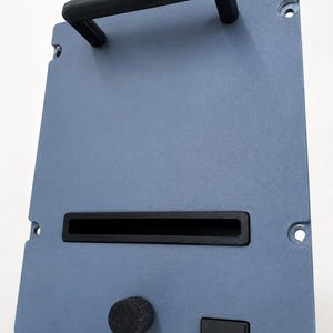 May include: A rectangular blue metal panel featuring a black top handle, a horizontal black slot in the center, a circular black knob, and a small rectangular black button on the lower section. The panel includes four mounting holes at the corners.