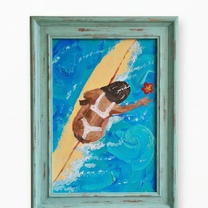May include: An acrylic painting titled Surfer Girl, Art by Adeline, featuring a figure in a white swimsuit on a yellow surfboard in blue ocean waves with a red flower. The artwork is enclosed in a distressed light blue wooden frame.