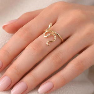 Könnte beinhalten: Ein goldfarbener Ring mit einem schlanken, gewundenen Schlangendesign, das sich um den Finger legt. Das Metall hat eine polierte, reflektierende Oberfläche, und der Schlangenkopf befindet sich oben, wobei der Körper ein offenes Band bildet.