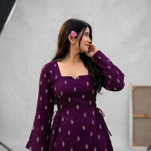 May include: A deep purple long-sleeved dress featuring a square neckline and a repeating white floral pattern. The garment includes side ties at the waist and long, slightly flared sleeves, providing a structured yet fluid silhouette.