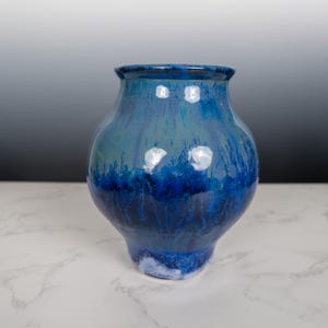 May include: A handcrafted ceramic vase with a glossy, blue glaze. The vase features a rounded body and a slightly flared rim. The color transitions from light blue at the top to a deep, dark blue at the base, creating a striking visual effect. The vase is suitable for home decor.