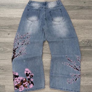 Könnte beinhalten: Hellblaue Jeans aus Denim mit weitem Bein und hoher Taille. Die Rückseite der Jeans ist mit gestickten Kirschblütenzweigen in Rosa-, Weiß- und Dunkelbrauntönen verziert, die sich vom Saum aus nach oben über die Hosenbeine erstrecken.