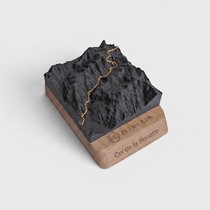 May include: A 3D topographic model of the Col de la Bonette mountain pass featuring a black terrain surface with a gold path line. The base is made of light brown wood and is engraved with the text HC, 23.3 km, 6.8%, and Col de la Bonette.