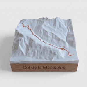 May include: A white 3D topographic map model of the Col de la Madeleine mountain pass, featuring a raised copper-colored path winding across the terrain. The model is mounted on a rectangular wooden base with the text Col de la Madeleine engraved.