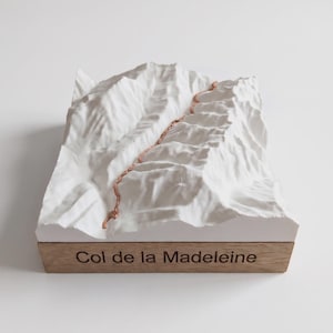 May include: A white 3D topographic model of the Col de la Madeleine mountain pass featuring a textured surface and a copper-colored path winding through the peaks, mounted on a rectangular wooden base with the text Col de la Madeleine.
