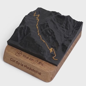 May include: A 3D topographic model of the Col de la Madeleine mountain pass. The black terrain features a gold-colored winding road path. The wooden base is engraved with the text HC, 19.6 km, 7.8%, and Col de la Madeleine.