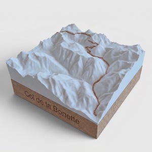 May include: A 3D topographic model of the Col de la Bonette mountain pass. The white terrain features a raised copper-colored path winding through the peaks. The model is mounted on a square wooden base with the text Col de la Bonette engraved.