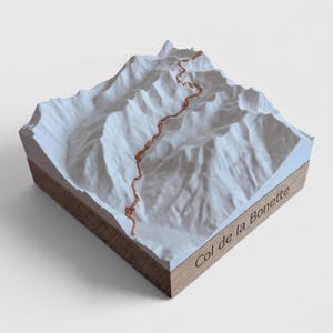 May include: A 3D topographic model of the Col de la Bonette mountain pass. The white terrain features a winding copper-colored road path across the peaks. The model is mounted on a square wooden base with the text Col de la Bonette engraved.