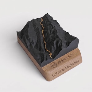 May include: A 3D topographic model of the Col de la Madeleine mountain pass, featuring a black matte terrain with a gold path. The base is made of light brown wood, engraved with the text "HC 25.9 km 6.2%" and "Col de la Madeleine".