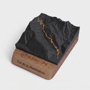 May include: A 3D topographic model of the Col de la Madeleine mountain pass featuring a black textured terrain and a gold path. The wooden base is engraved with the text HC 19.3 km 7.9% and Col de la Madeleine in a dark brown font.