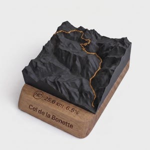 May include: A 3D topographic model of the Col de la Bonette mountain pass featuring a black terrain surface with a gold-colored path. The base is made of light brown wood and is engraved with the text HC, 25.6 km, 6.5%, and Col de la Bonette.