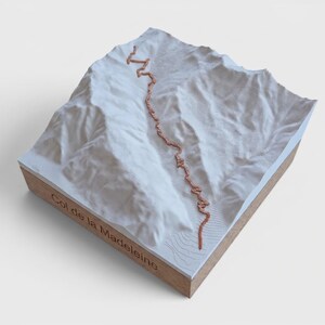 May include: A 3D topographic map of Col de la Madeleine featuring a white mountain relief on a wooden base. A copper-colored line traces the winding path across the terrain. The text Col de la Madeleine is engraved on the side of the wooden base.