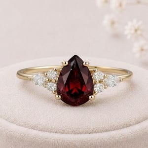 May include: A gold ring featuring a central pear-shaped deep red garnet gemstone. The band is accented on both sides by a cluster of three small, round, clear white stones, creating a symmetrical design that highlights the dark red center.