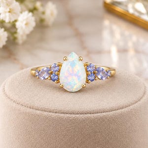 May include: A gold ring featuring a central pear-shaped opal gemstone flanked by clusters of small, light blue marquise and round-cut stones. The setting is polished gold, showcasing the iridescent play of color within the opal and the cool blue tones of the accent gems.