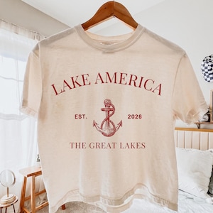 Peut inclure: Un t-shirt &agrave; manches courtes de couleur cr&egrave;me avec un texte rouge indiquant LAKE AMERICA, EST. 2026 et THE GREAT LAKES. Un graphique rouge repr&eacute;sentant une ancre entour&eacute;e d'une corde est centr&eacute; entre le texte. Le t-shirt est sur un cintre.