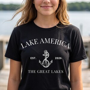 Peut inclure: Un t-shirt noir &agrave; manches courtes avec le texte blanc LAKE AMERICA, EST. 2026 et THE GREAT LAKES. Un graphique blanc d&eacute;taill&eacute; repr&eacute;sentant une ancre marine entour&eacute;e d'une corde est centr&eacute; entre la date de cr&eacute;ation et le texte.