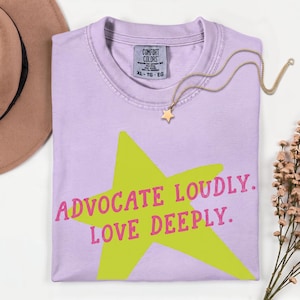 Könnte beinhalten: Ein hellviolettes T-Shirt mit einem großen, lindgrünen Stern-Grafikmotiv in der Mitte der Brust. Der Text "ADVOCATE LOUDLY. LOVE DEEPLY." ist in Pink über den Stern gedruckt. Das T-Shirt hat einen Rundhalsausschnitt und ein Comfort Colors Etikett.
