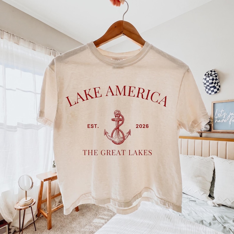 Peut inclure: Un t-shirt &agrave; manches courtes de couleur cr&egrave;me avec un texte rouge indiquant LAKE AMERICA, EST. 2026 et THE GREAT LAKES. Un graphique rouge repr&eacute;sentant une ancre entour&eacute;e d'une corde est centr&eacute; entre le texte. Le t-shirt est sur un cintre.