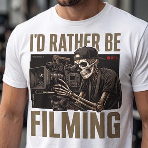 Op de afbeelding: Een wit t-shirt met een afbeelding van een skelet met een zwarte honkbalpet die een professionele videocamera bedient. De tekst "I'D RATHER BE FILMING" staat in grote bruine letters boven en onder de centrale illustratie gedrukt.