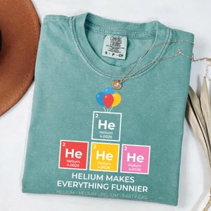 Puede incluir: Una camiseta de manga corta color verde salvia con un gráfico de tres globos coloridos sobre cuatro cuadrados de la tabla periódica para el Helio. El texto dice "He" con "Helium 4.0026" y la frase "HELIUM MAKES EVERYTHING FUNNIER" con "HELIUM" repetido.