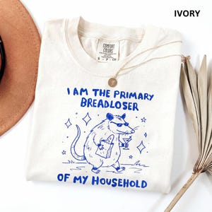 May include: An ivory t-shirt featuring a blue graphic of an opossum wearing sunglasses, holding a paper bag and a martini glass. The text reads: I AM THE PRIMARY BREADLOSER OF MY HOUSEHOLD. The shirt has a Comfort Colors brand label.
