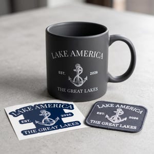 Peut inclure: Un mug en c&eacute;ramique noir mat avec le texte blanc LAKE AMERICA EST. 2026 THE GREAT LAKES et un motif d'ancre central. &Agrave; c&ocirc;t&eacute; se trouvent un autocollant carr&eacute; assorti et un &eacute;cusson en tissu rectangulaire, arborant tous le m&ecirc;me motif.