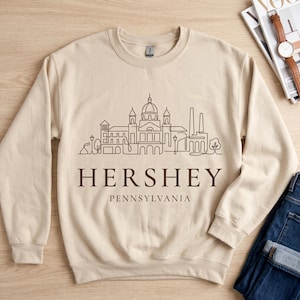 Może przedstawiać: Piaskowa bluza z okrągłym dekoltem, ozdobiona ciemnobrązową grafiką liniową przedstawiającą panoramę Hershey w Pensylwanii. Napis HERSHEY PENNSYLVANIA został nadrukowany ciemnobrązową czcionką szeryfową pod ilustracją architektoniczną.