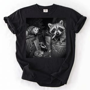 Może przedstawiać: Czarny t-shirt z czarno-białym nadrukiem graficznym przedstawiającym szopa pracza trzymającego papierosa na metalowym śmietniku, ze skunksem stojącym przy worku na śmieci i opossem siedzącym na drewnianym płocie w tle.