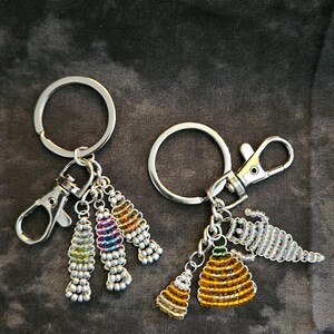 May include: Two silver-toned metal keychains featuring lobster claw clasps and split rings. One keychain holds three beaded charms in silver, pink, blue, and orange. The other keychain features a beaded beehive, a cone, and a silver-toned lizard charm.