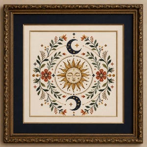 May include: A framed cross-stitch embroidery featuring a central sun with a human face, surrounded by two crescent moons, floral motifs, and leafy branches in shades of gold, dark blue, green, and rust red on a cream-coloured fabric background.