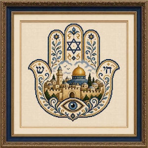May include: A framed cross-stitch embroidery depicting a hamsa hand symbol. The design features a central cityscape with a golden dome, a Star of David at the top, and a blue eye at the bottom, all rendered in blue, gold, and beige thread colors.