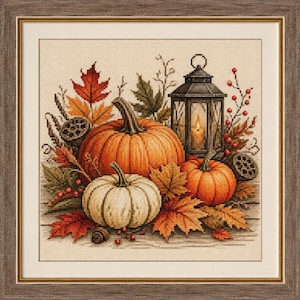 May include: A framed cross-stitch embroidery featuring two orange pumpkins and one white pumpkin surrounded by autumn leaves, red berries, and a black lantern with a glowing candle. The artwork is set within a textured brown wooden frame.