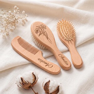 Könnte beinhalten: Ein Set aus drei hellen Holzkämmen und Bürsten, bestehend aus einem Kamm, einer weichen Haarbürste und einer Bürste. Der Kamm und der Bürstengriff sind mit dem Namen Amelia graviert, und der Bürstenkopf zeigt eine florale Strichzeichnung.